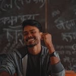 Young South Asian student smiling in front of a chalkboard in a Bangladeshi classroom.